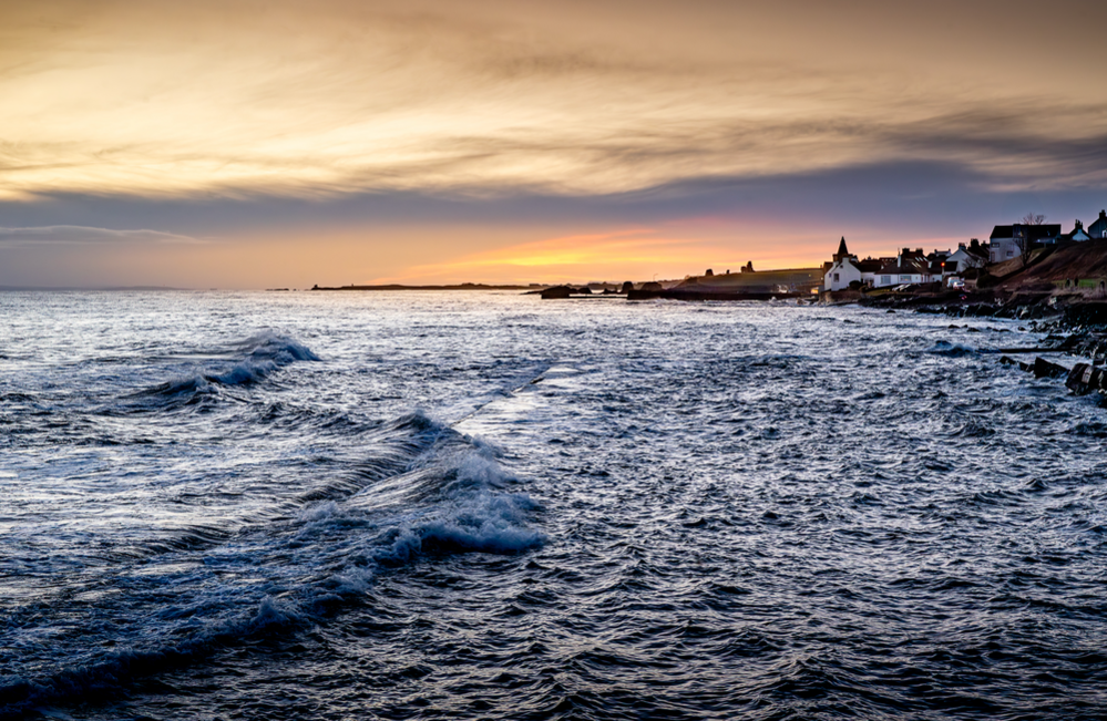 A sunset over the water. Waves lap the shore and a small village can be seen in the bay.