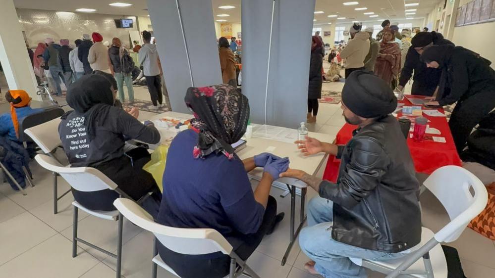 The images shows a wide view of the blood donation event held at the Guru Nanak Gurdwara in Luton on Sunday. In the foreground organisers and staff at the event are sat with people at tables offering finger prick blood tests. In the distance a line of people can be seen queuing. 