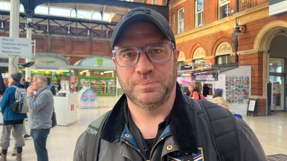 Ian Tuthill pictured in a black jacket and black shirt, wearing blue rimmed glasses and a baseball cap. He is standing on the concourse at Norwich railway station, with other people nearby, and the station Co-op and florist stall in the background.