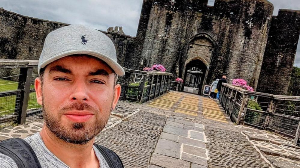 Bobbie Rammond takes a selfie. He is wearing a grey baseball cap and has dark facial hair. Behind him is the entrance to a castle.