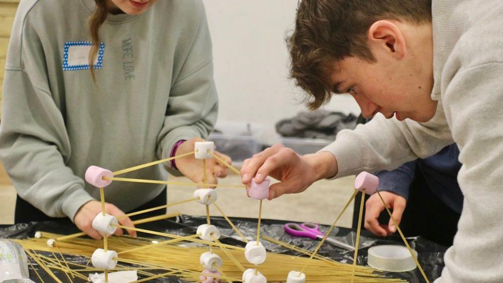 Young people studying a challenge on a table with marshmallows and spaghetti.