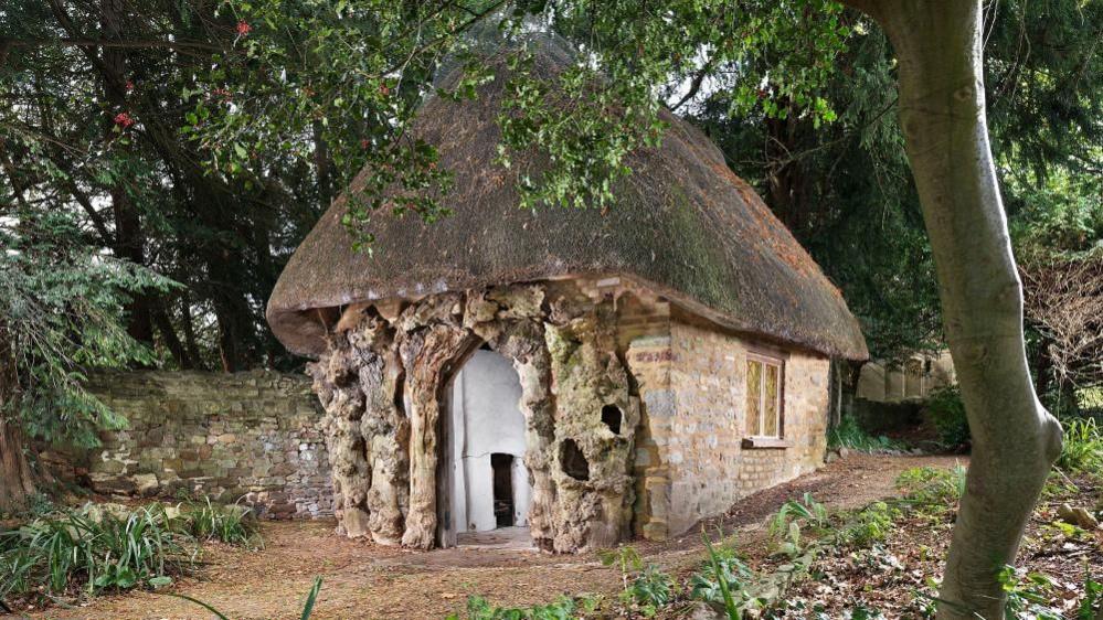 General view of brick and rubblestone hut with thatched roof and front wall of applied rustic tree trunks framing entrance.