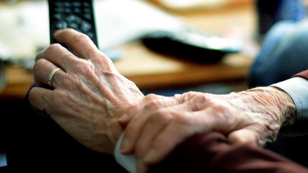 An older persons hands seen holding a TV remote control. There is a wedding ring on the left hand and the ends of the arms are covered by a light blue shirt and a burgundy jumper.