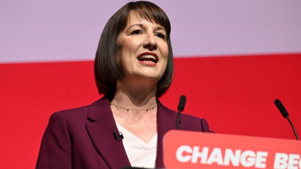 Rachel Reeves standing at a lectern with a maroon suit and white top 