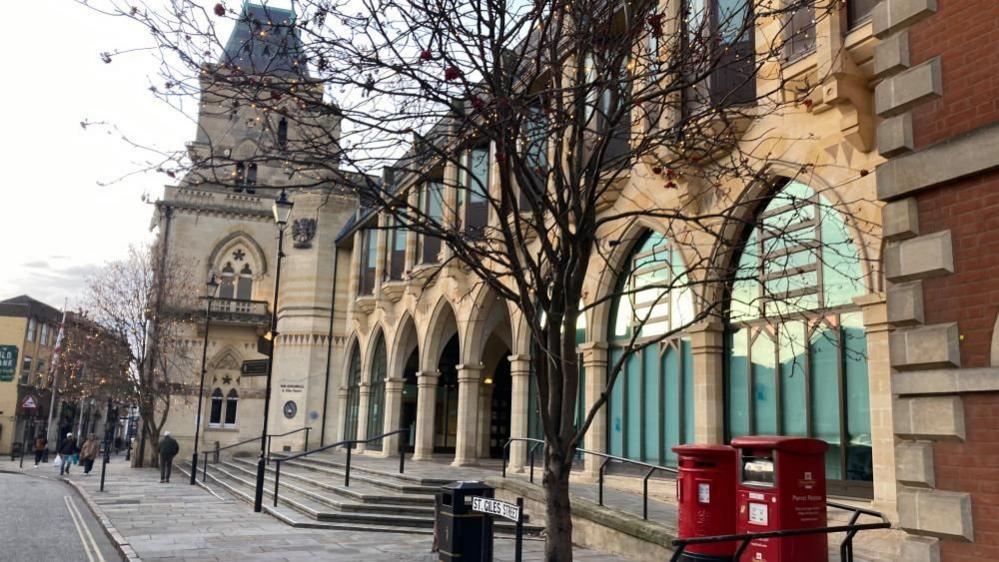 The Guildhall in Northampton, showing two parts of the building. One with more modern arches and another building, to the right that is more historical. There are two red post box boxes to the right, a black bin and a street sign. There are trees outside the buildings and people are walking past it in the distance. 