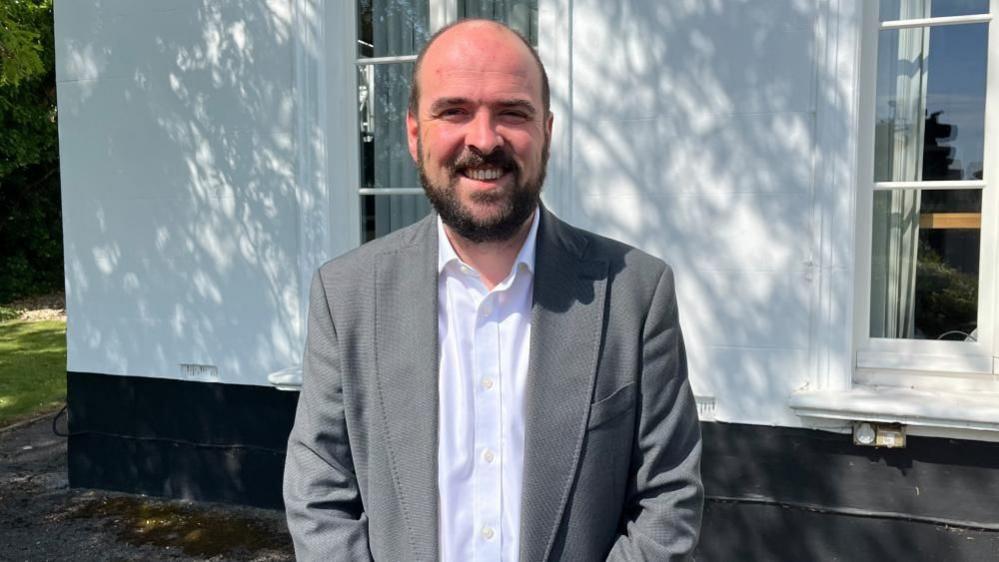 Richard Holden standing in front of a white-walled building. He has a beard and is smiling. He is wearing a grey jacket and white shirt.