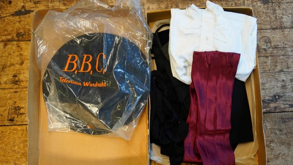 In a box, a white filled shirt, silk red fabric and what appears to be a black hat embroidered in orange reading BBC television wardrobe