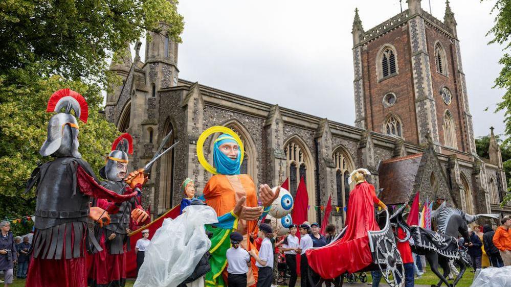 St Albans cathedral with a procession of people walking past, some wearing huge puppet costumes -Roman centurions, St Alban himself and a chariot. A group of children in scout hats and white t shirts stand around the saint puppet.