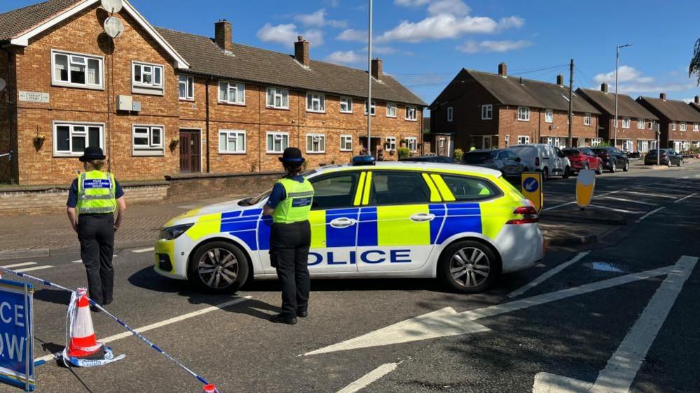 Two police officers standing in a street, by a police car, with their back to the camera