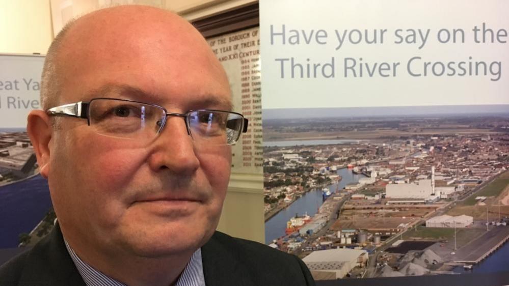 Councillor Graham Plant, standing next to a banner depicting plans for the Third River Crossing, which was subsequently named Herring Bridge. He has close cropped hair and is wearing glasses, and a suit with blue and white striped shirt. He is pictured inside Great Yarmouth Town Hall and over his left shoulder is one of several marble tablets within the reception listing the aldermen and mayors of the borough from 1208 to present day.