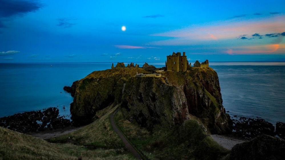 An evening snap of Dunnottar Castle near Stonehaven. The sky is blue and it's getting darker. A ruined castle stands atop a small and tall patch of land surrounded by water with a little path leads back to the mainland, where the photographer is. The moon is out and the sky is streaked with orange.