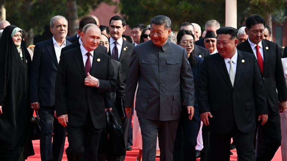 Putin, Xi and Kim walking along smiling, with many people in their entourage behind them.