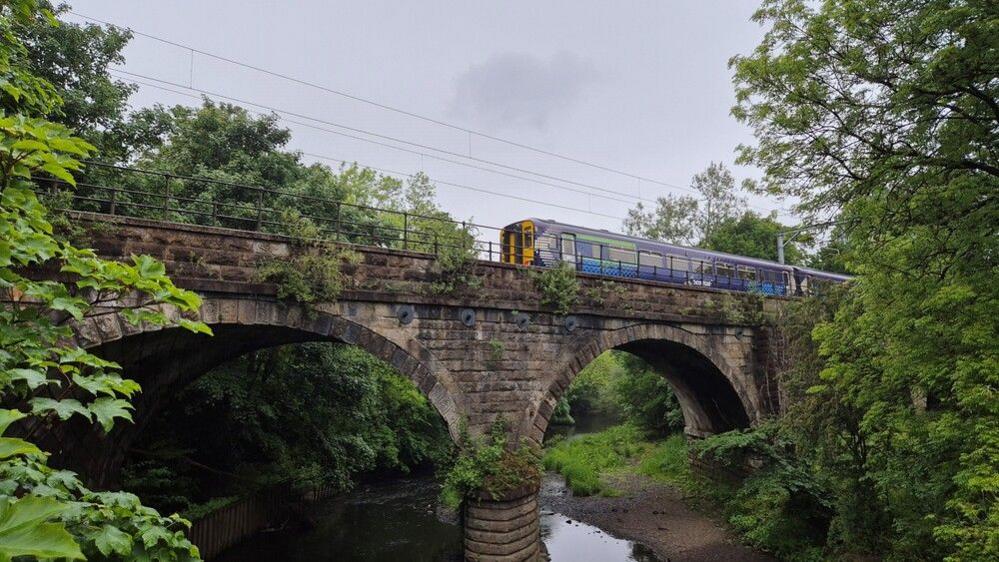 A ScotRail train crosses over a bridge with two archways over a river. It is surrounded by trees on both sides.