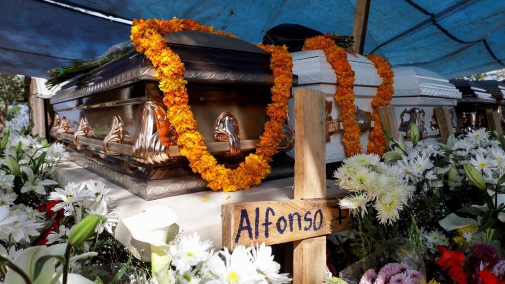 A row of caskets lined up at a wake for a family murdered in Chilpancingo. The caskets are covered in flower garlands and a cross placed in front of one of them bears the name "Alfonso" and the number seven, the age of the boy who was among those killed. 