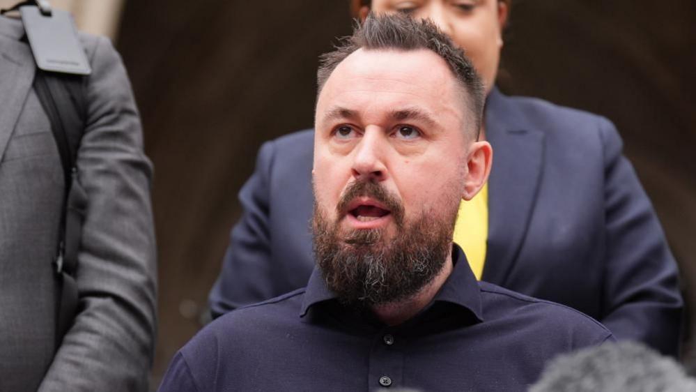 Martin Hibbert sits speaking. A media microphone is in front of him and people stand behind him.