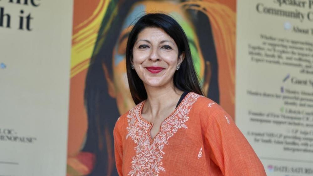 Sheetal Jethwa, founder of South Asian Voices Bristol CIC, poses for a picture in front of large posters. She is wearing an orange top with white embroidery around the neckline and red lipstick. She has long, dark hair.