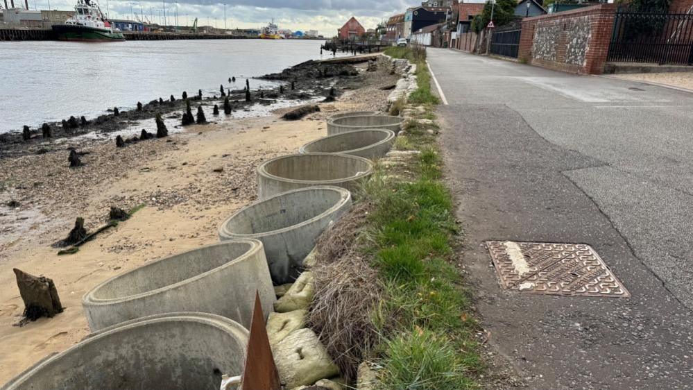 A series of concrete pipes, listing and lilting owing to the impact of tide. They are no longer against the edge of Riverside Road, which runs to the right of this image. The river is on the left.