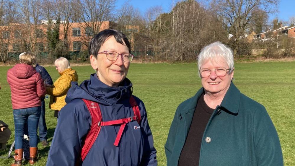 Judith Morris with short dark hair and glasses wearing a blue raincoat with red straps, and Liz Scott with short white hair and glasses, wearing a green coat and black top. They are standing in a grass field with trees behind.  A small group of people is also visible to the left.