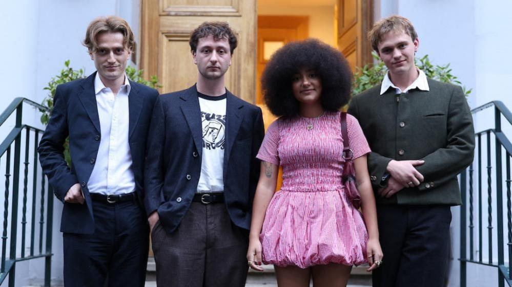 English Teacher band members Nicholas Eden, Lewis Whiting, Lily Fontaine and Douglas Frost standing on steps in front of an open door.