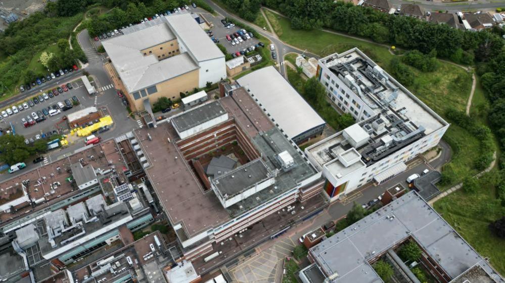 Aerial shot of Kettering General Hospital, showing three square buildings and an l-shaped block.  There are a number of small car parks nearby.