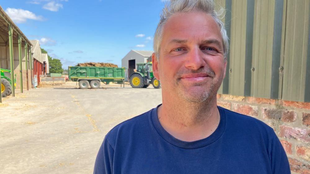 A man wearing a blue T-shirt smiles at the camera. He is standing in a farmyard with a tractor in the background pulling a trailer full of a brown substance.