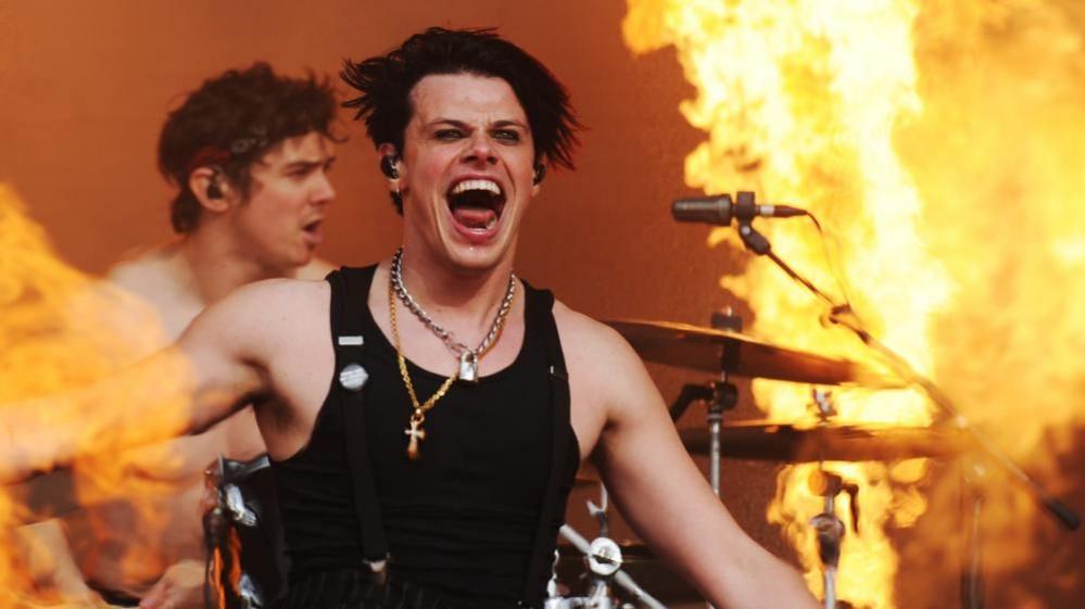 Yungblud performs in front of a drummer and flames, used for effect on the stage, at a festival.
