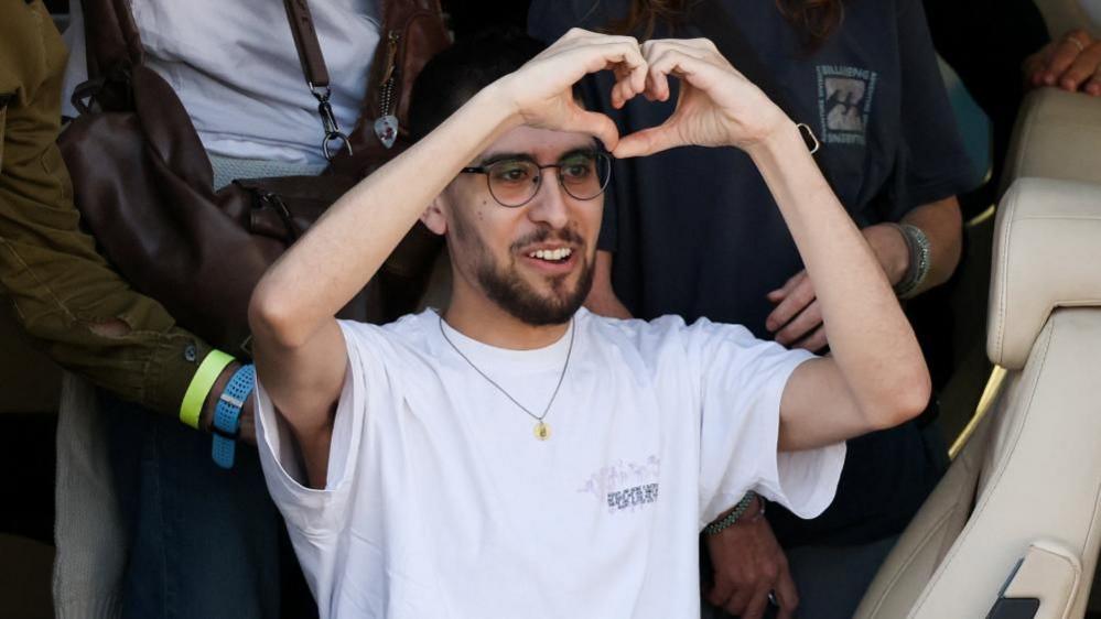 Evyatar David, wearing a white T-shirt, a necklace, glasses and sporting a beard, makes the sign of a heart with his hands just above his head