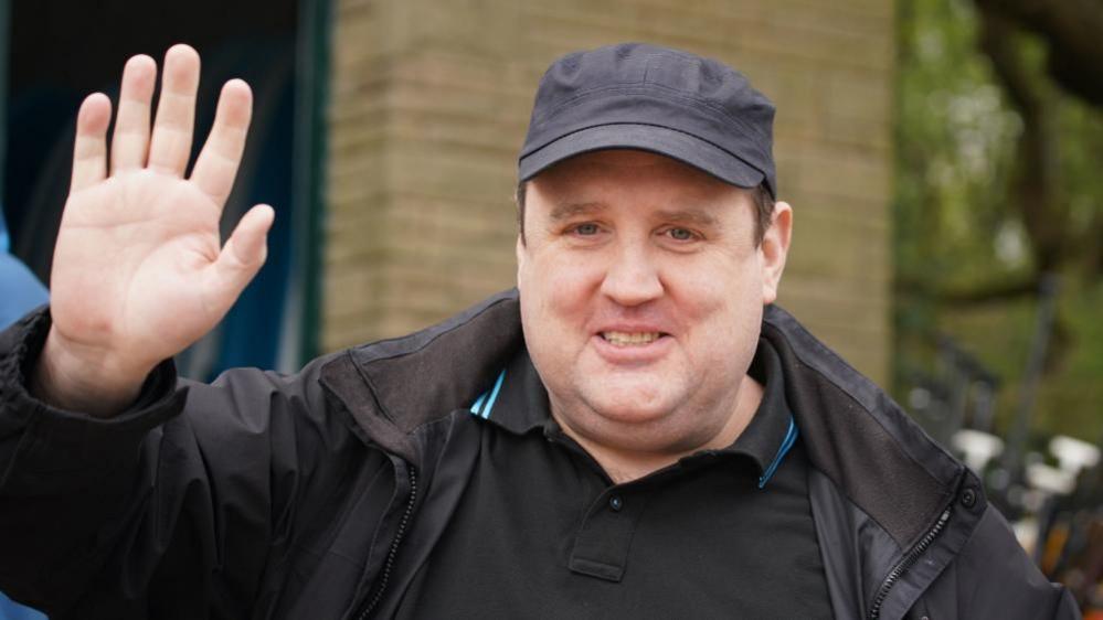 Peter Kay stands in a street wearing a black jacket and cap. He is waving at the camera. 