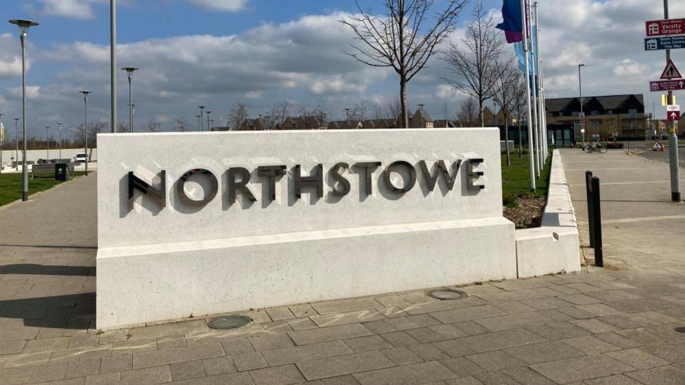 A sign that says Northstowe. It is a white concrete plinth with the name written in metal. A building is in the background to the right. There are signs on a lamp post and trees can be seen in the distance behind the plinth. There is a row of flags on the right.
