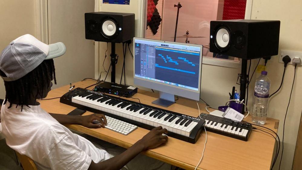 A teenager wearing a silver and white baseball cap and white T-Shirt, is sat at a computer with two keyboards and speakers on a wooden desk in front of him.