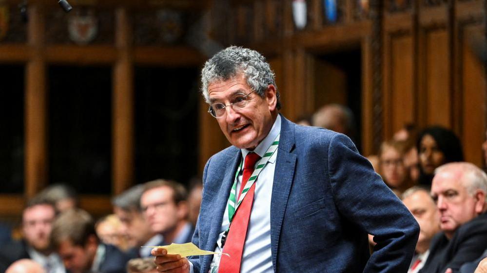 A man standing in the House of Commons, holding a note and speaking to the House. He is wearing a blue suit and a red tie, and he has glasses on