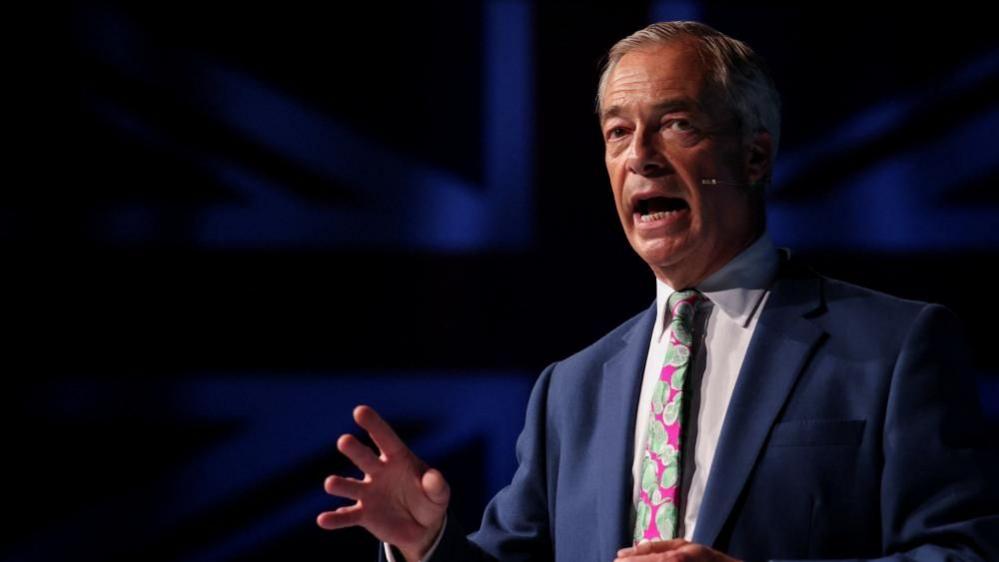 Nigel Farage, wearing a pink and green patterned tie is pictured mid-speech with a dark background with the union flag faintly visible behind him. 