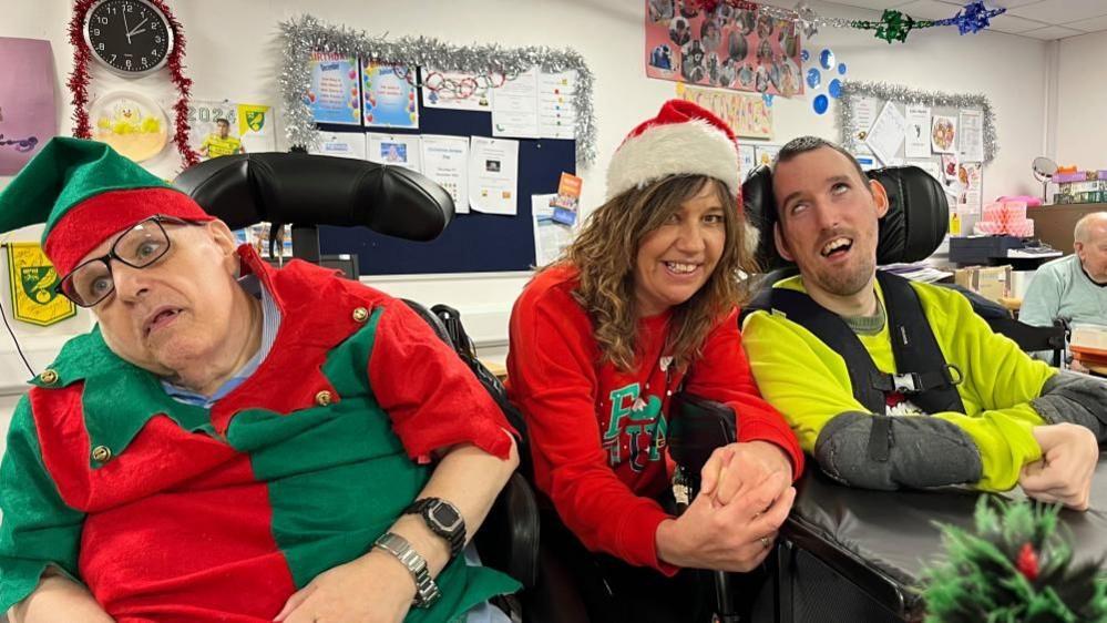 Julie Charles, wearing a Santa hat and jersey, has long auburn hair. SHe is smiling and holding her hands together. She is sat between two of the members of Centre81; the gentleman on the left is wearing a red and green elf outfit, and a wrist watch and glasses. The man on the right has close cropped hair, and is wearing a high visibility jersey, and has a chest harness and elbow pads.