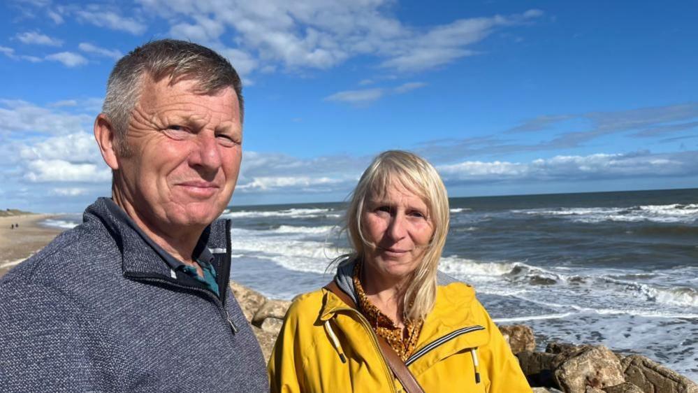 David Loades in a blue top and Amanda Loades in a yellow jacket, standing near the sea and coastal defences