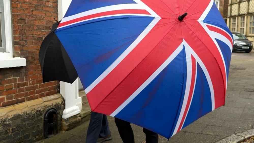 Two men hidden behind umbrellas outside King's Lynn Crown Court. One umbrella is black, covering Mr Harris' face; the other is a Union Flag pattern, covering another man accompanying Mr Harris.