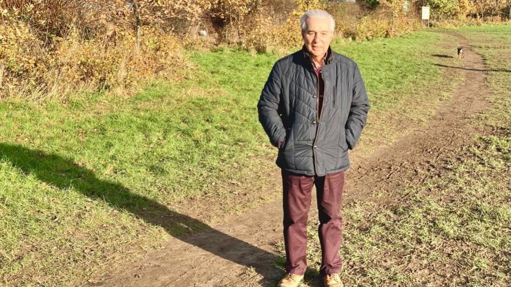 Grey-haired man wearing blue coat, red trousers and brown shoes standing in field with hands in  pocket. His shadow falls to the right as we look and a path runs behind him. A dog can be seen on the path.