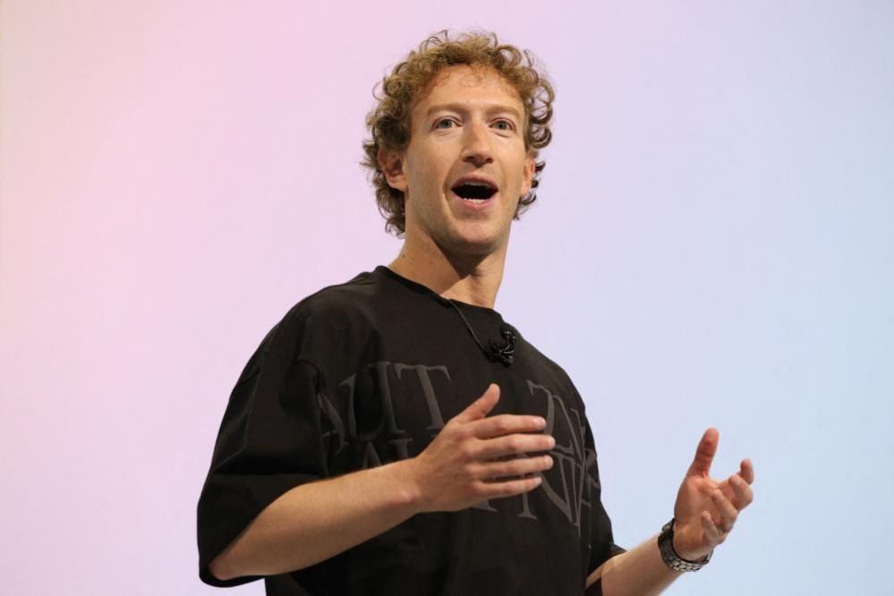 Meta chief Mark Zuckerberg seen on stage during a speech. He has colour ginger hair and is wearing a baggy black t-shirt. He is holding his hands in front of him as he speaks. 