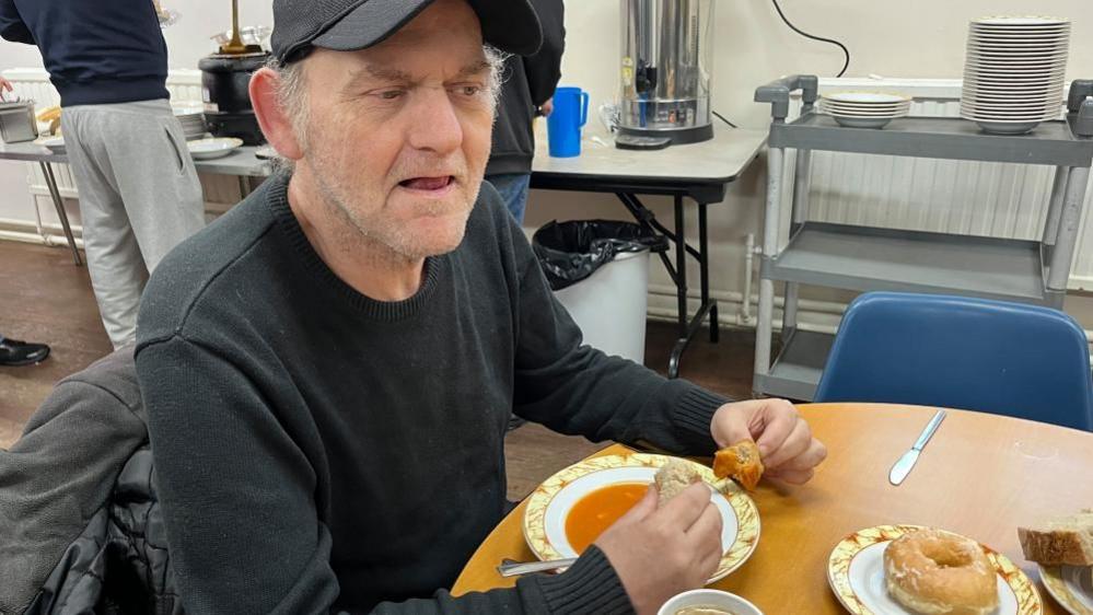 Michael Lucas with blue cap sits with a bowl of soup