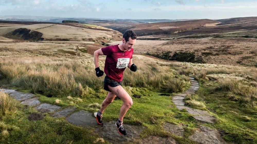 A man runs over green, muddy hills, wearing black shorts a red T shirt and a name label. 