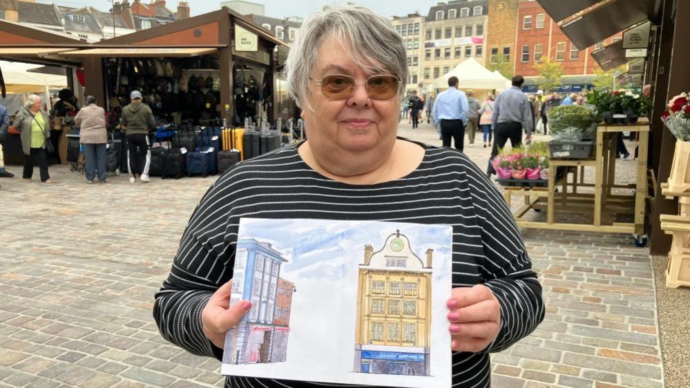 Rosie with medium-length grey hair and darkened glasses wearing a black and white top in the Market Square. She is holding paintings of two buildings in the square.