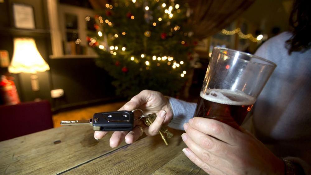 A pair of woman's hands holding an alcoholic drink in one, and car keys in the other with a lit up Christmas tree in the background.