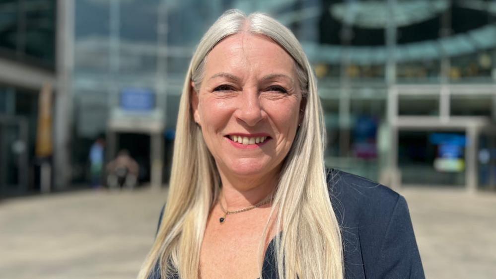 Kay Mason Billig is smiling. She has long blonde hair and is wearing a dark jacket. She is standing outside The Forum in Norwich, which appears in soft focus.