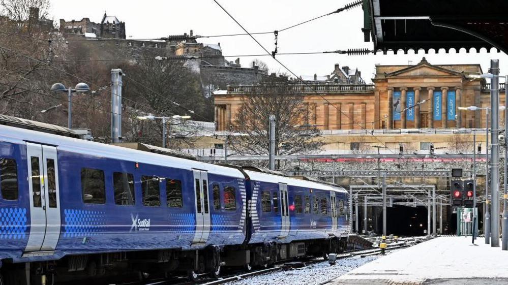 ScotRail train at Edinburgh's Waverley station. There is snow on the platform. The National Galleries of Scotland overlooks the track and Edinburgh Castle is on the hill above