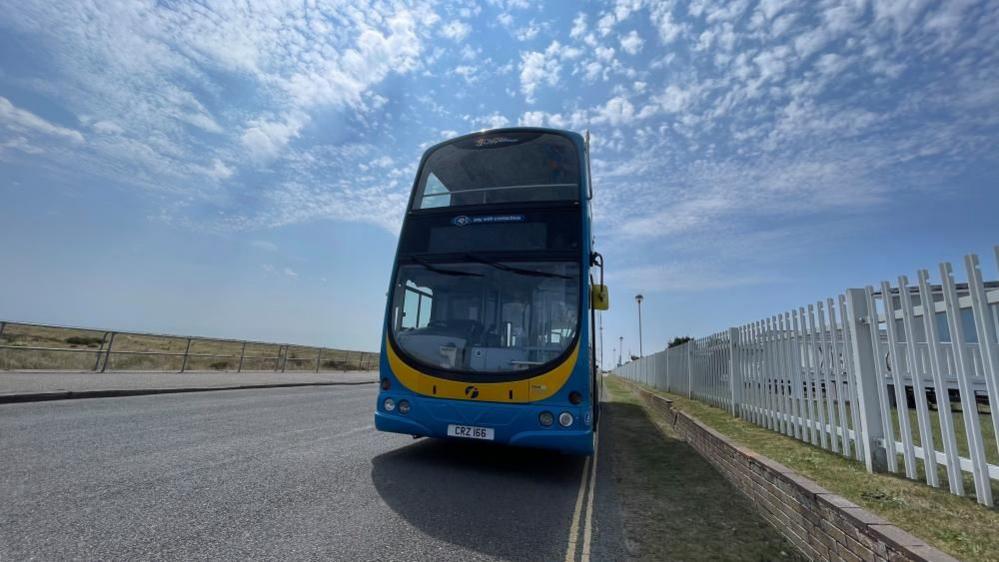 The Coastal Clipper Cabriolet is a blue bus, parked on Great Yarmouth sea front next to the Haven Seashore Holiday Park with picket fencing to the right of the image and the beach to the left. A mackerel sky puts the vehicle in silhouette!