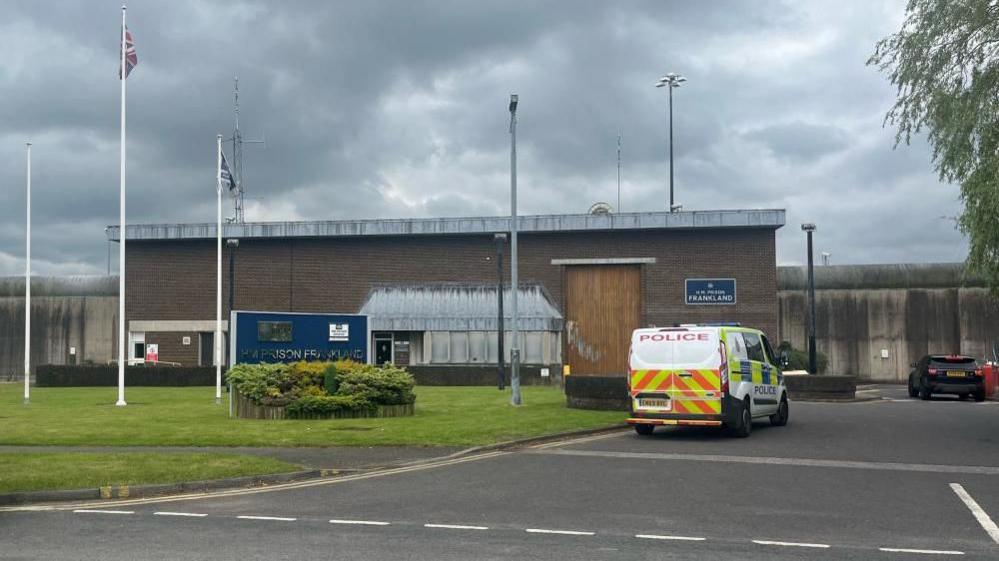 A police van is parked outside a low flat-roofed building with a plaque reading 'HMP Frankland' on the wall