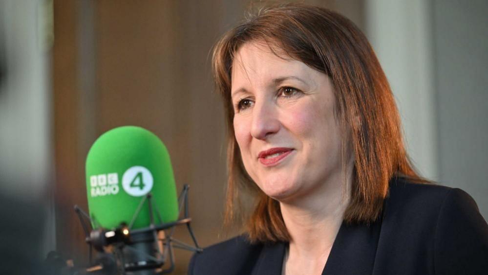 A woman with brown hair and wearing a black jacket vaguely smiles as she speaks into microphone.