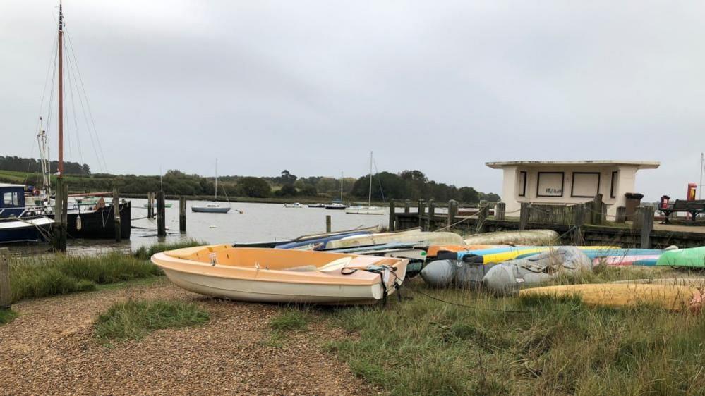 A yellow and white rowing boat, in front of a variety of other boats, resting on a grass and gravel area near a river