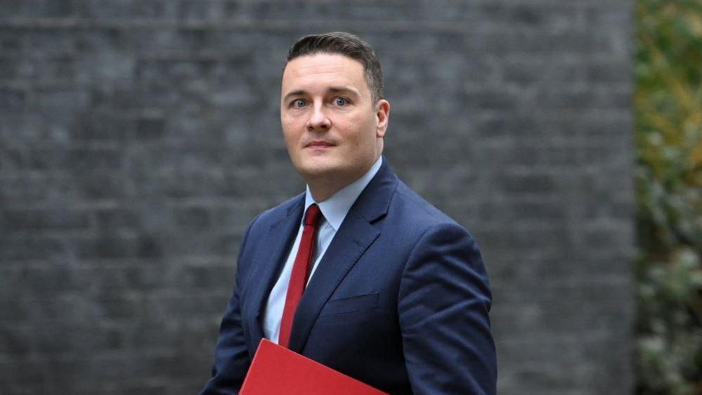 Wes Streeting walks outside Downing Street. He has short brown hair and is wearing a blue suit. He is holding a bright red folder.