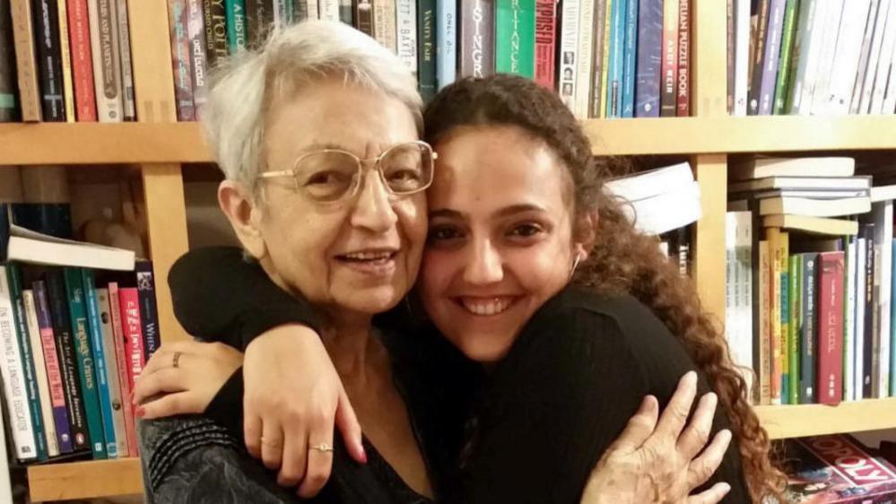 Family handout photo of Romi Gonen hugging her grandmother. Both are embracing and smiling at the camera, infront of a bookshelf