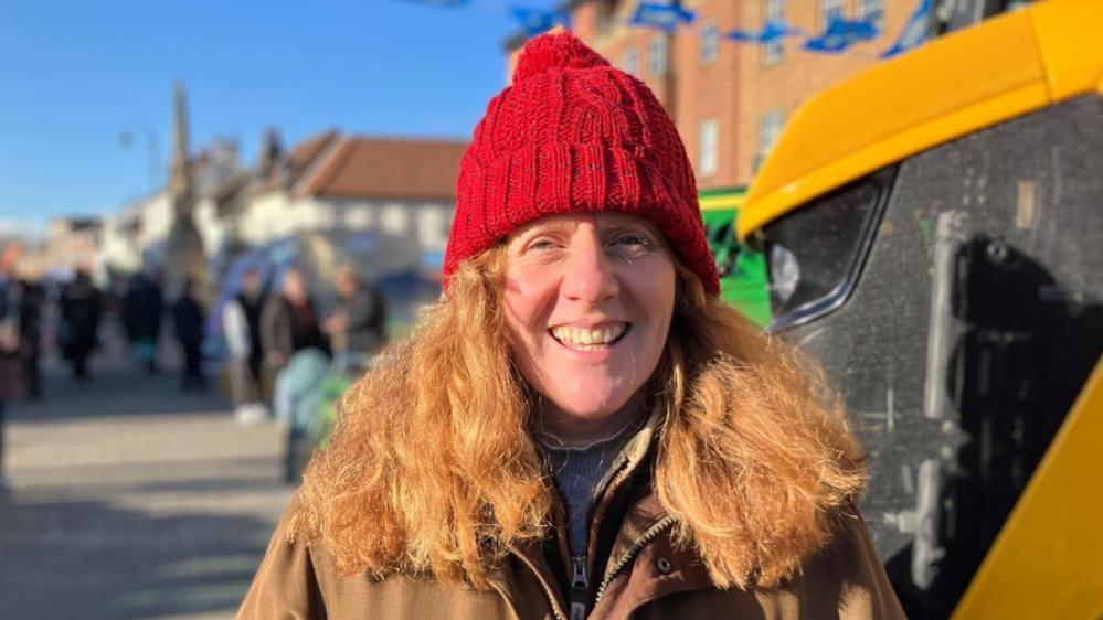 Sophie Bramley is smiling looking straight ahead. She has long hair and is wearing a red hat with a brown jacket. She is stood at the side of a yellow tractor. 